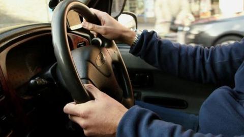 A close up shot of a set of male hands on a car's steering wheel. The man is wearing a long sleeved blue top and a watch on his right hand