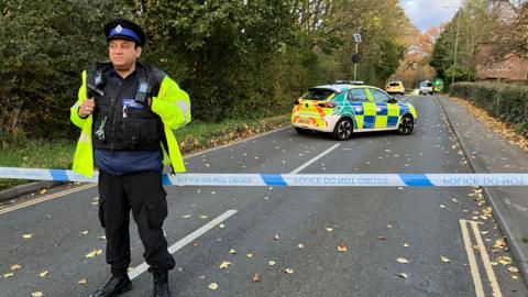 Police officer in front of a cordon tape across a main road, with several police vehicles in the background.