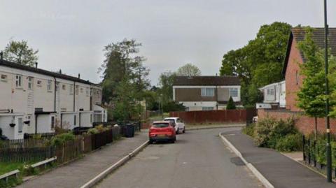 A general view of the street with white-painted terraced houses on the left and newer red-brick properties on the right. There are trees in the background and cars parked on the street. 