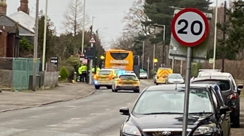 A view from several feet away of a yellow bus parked by the side of a road, with police officers standing beside it, and two police cars parked behind it