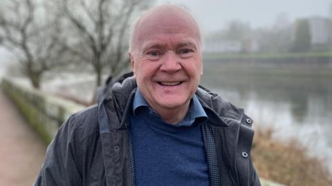 A grey haired, balding man with a dark jacket and blue jumper and shirt stands in front of the River Nith in Dumfries