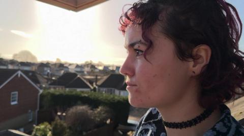 Niamh Tickner, a woman in her twenties with short, curly purple hair. She has an eyebrow piercing and a piercing in her nose. She is wearing a dark blue paisley top and a black choker. In the background is a blurry cul-de-sac of houses.