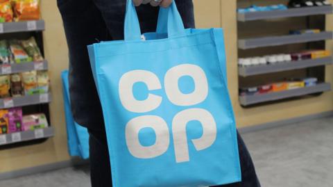 A shopper carrying a blue Co-op bag inside a store. There is an array of products on a beige wooden shelf in the background.