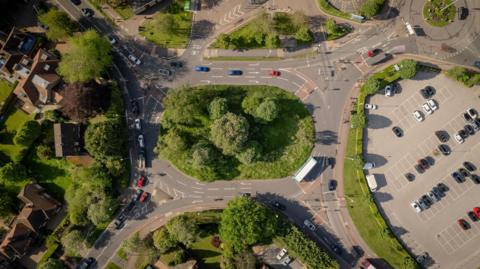An aerial view of a roundabout with four exits and green bushes and trees in the centre.