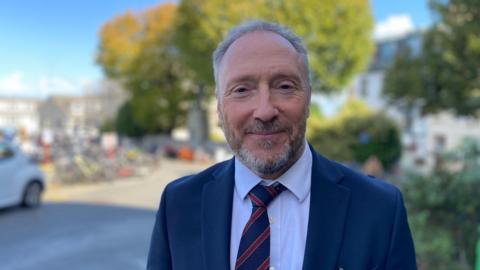 Image shows Deputy of the Committee for Education, Sport & Culture, Paul Montague. He wears a navy blue suit, with white shirt and blue and red-striped tie, with grey hair and beard, smiling into the camera, against a backdrop of a sunny day, blue sky, green trees and houses/ buildings, blurred out.