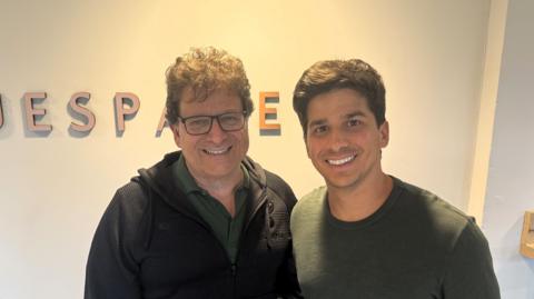 A father and son, both with dark hair, pose for the camera in front of a white wall