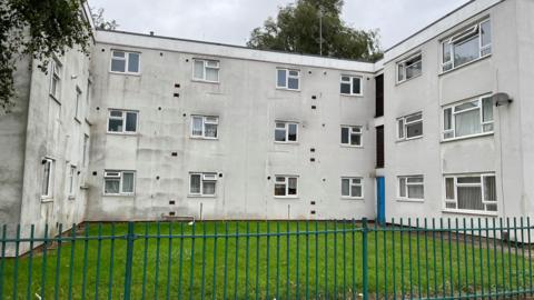 White block of flats at Alliston gardens with a green fence around the edge and grass in front
