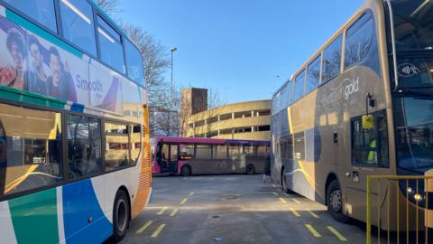 Two parked double-decker buses parked in a bus station while a single decker purple bus drives away.