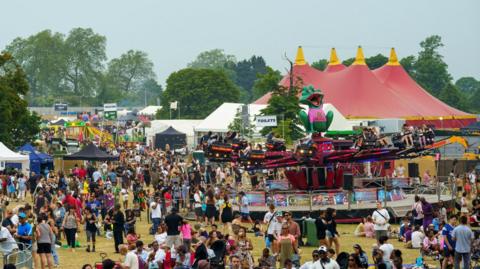 A busy scene is depicted from the Lambeth Country Show 2024. It shows crowds of people wearing summer clothes, and fairground rides, as well as white event tents.