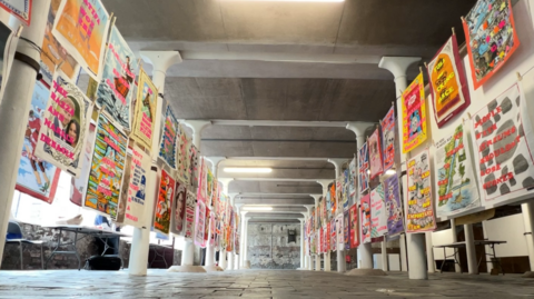 Two rows of bright tea towels are hung on string on either side of a large space. The space has lots of white pillars holding up a concrete ceiling. The tea towels have large messages, which can't quite be read, and vivid images. 