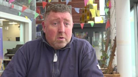 Emlyn Jones, a former teacher who runs a security firm. Mr Jones sits in front flags in a school building. He is wearing a navy sweatshirt quarter zip. 