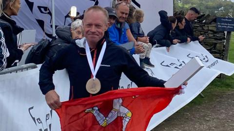 Geoff, a man with short blond hair and wearing a large medal, smiles and holds up a Manx flag.