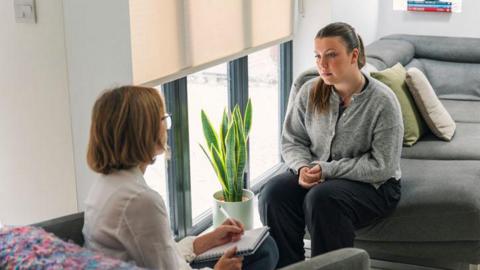 A patient sits on a sofa speaking to a clinician taking notes during a consultation in a room with large windows and a houseplant.