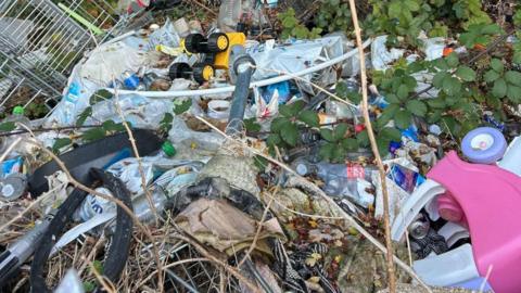A pile of rubbish on grass. Included in the rubbish are empty plastic bottles, a trolley, a plastic children's car and carrier bags.