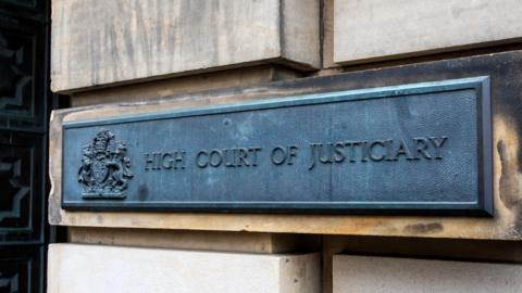 A grey metal sign on the sandstone block wall of the High Court in Edinburgh. The sign has a royal crest and reads: "High Court of Justiciary". 