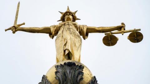 A golden "scales of justice" statue showing a woman wearing robes and a crown with her arms outstretched. In one hand she holds a sword pointing upwards, in the other she holds a set of scales. The sky is grey.