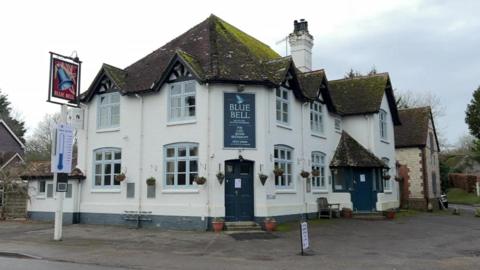 A white pub with a blue sign on the front which says "Blue Bell" on it. There is also a blue cartoon thermometer on a sign showing how much money has been raised.