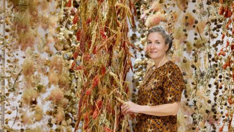 Artist Rebecca Louise Law standing beside red and yellow flowers suspended from the ceiling, Her grey hair is pulled back and she is wearing an orange top. She has blue eyes and is smiling slightly at the camera. 