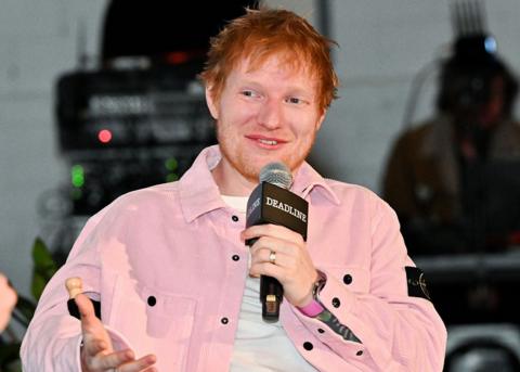 Ed Sheeran speaks onstage while wearing a pink cord jacket 