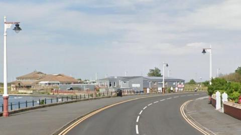 The Promenade in Southport - a road winds to the right - lined with double yellow lines. On the left is water and some grey buildings. on the right is gates into a residential home with green bushes. 