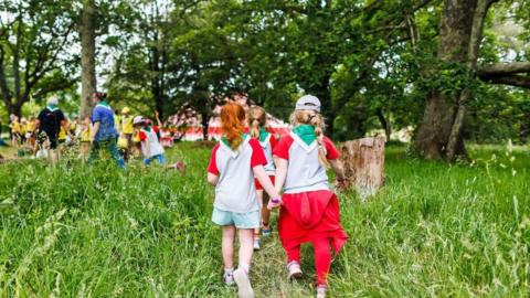 A close-up of two young girls walking hand-in-hand through long grass - one has ginger hair and the other blonde, they wear red and white t-shirts.