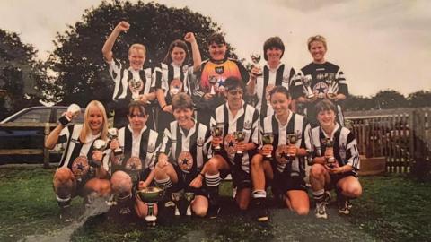 Newcastle Women FC women, wearing black and white striped football shorts, and black shorts and football boots, crouch down and some stand up, holding trophies on a grass field. 