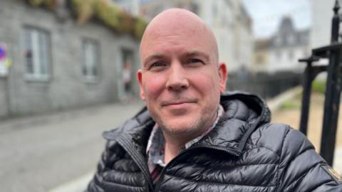 Marcus Saul is smiling at the camera as he sits outdoors. Blurred in the background behind him are stone buildings and a road. There are also thin black iron posts just behind him which may be part of the bench he is sitting on. Mr Saul is wearing a black puffer jackets and he has a shaven head, slight beard and blue eyes.