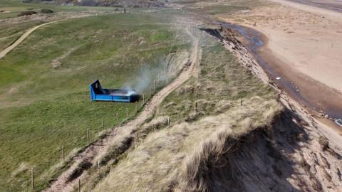 Smoke coming from small blue cordoned off structure on a grassy beach cliff. On the right hand side of the photo you can see sand.