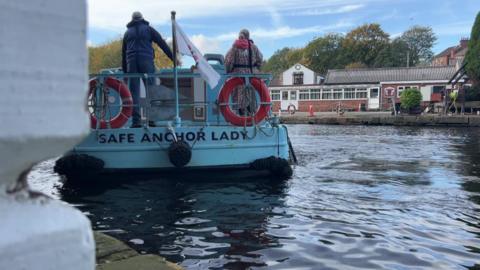 A picture of the back of one of then Safe Anchor Trust's boats, it's light blue with red safety rings and a flags, it's being steered by a man in a blue jacket with another person stood next to him. It's on the canal in Mirfield, the charity's building is in the distance.