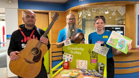 Denis Salgado from the School of Popular Music and Marcel Lenormand and Athene Sholl from Guernsey Art Network stood in the hospital. Denis is holding a guitar, while Marcel and Athene are stood behind a trolly full  or art supplies. 