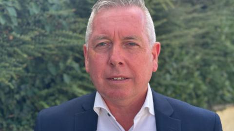 A man with short grey hair, a navy jacket and a white shirt.