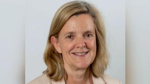 A woman with blonde, shoulder length hair in a white blazer smiles at the camera.
