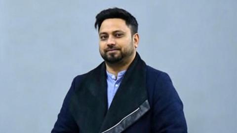 Masum Miah, wearing a dark blue coat, light blue shirt, and jeans, posing with his hands in his pockets against a neutral backdrop.