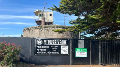 The circular brick fort is pictured behind black hoarding. It has a small wooden watchtower on top and a large tree to the right. On the black hoarding, which is preventing access, is a green "to let" sign and another black sign advertising it as a wedding venue.