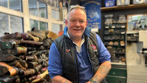 A photograph of a middle-aged man, smiling at the camera. He is white, with short light hair and tattoos on his arms, wearing a black gilet on top of a blue long-sleeved top. He sits in a shop, with shoes piled up in front of a window and tool shelving on the wall.