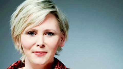 Michelle has short blonde hair and is wearing a red top with black leopard print on it, and two gold necklaces. 