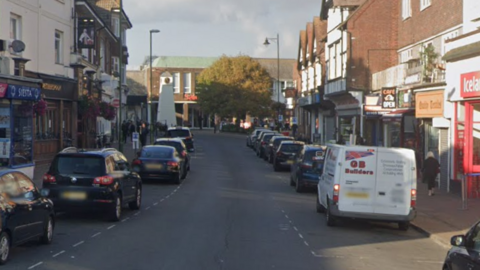 A Google Street image of a road with vehicles parked either side and shops either side.