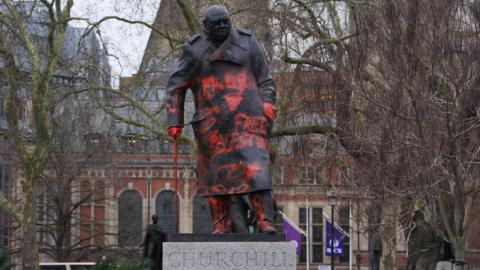 The bronze statue of Sir Winston Churchill in Parliament Square, London, is shown defaced with bright red spray paint. Large slogans, including "FREE PALESTINE," are visible across the front of the monument's coat. The red paint also covers the statue's hands and walking stick.