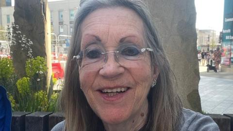 A selfie of Margaret Colling sitting outside in front of two tall decorative stones and planters. She is smiling and has long, straight greying hair and rim-less glasses