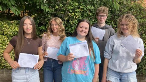 Young people get their results in Herefordshire, in a picture issued by the council. They are wearing summer clothes, holding pieces of paper and are looking happy.