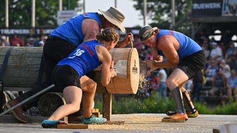 Two women brace themselves and crouch on either side of a huge wooden log while using a double-handled saw to slice through it