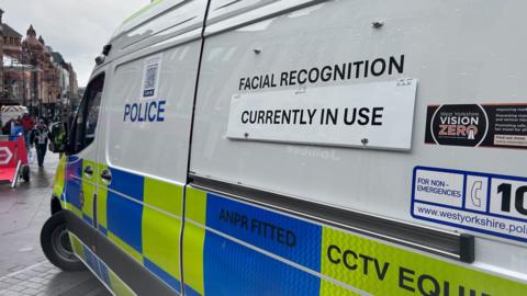 A close-up of a police CCTV van on Briggate in Leeds. A sign on the side of the van reads, "facial recognition currently in use". Shoppers can be seen in the background.
