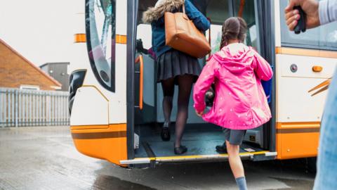 Stock image of two girls in school uniforms stepping on to a bus. One girl is also wearing a blue winter coat and a large brown handbag. The other girl has plaited hair and is wearing a pink coat with white polka dots.