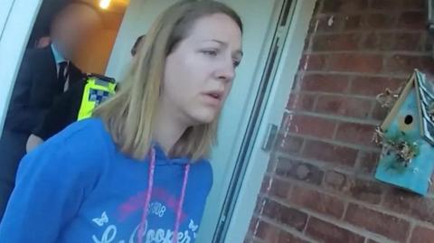 Police bodycam footage of Lucy Letby, with straight blonde hair, and wearing a blue hoodie, being led from her home with her arms behind her back