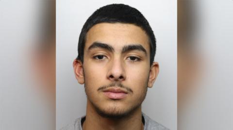 Tyler Wilkins with short dark hair and a slight beard wearing a grey top in a police custody photo