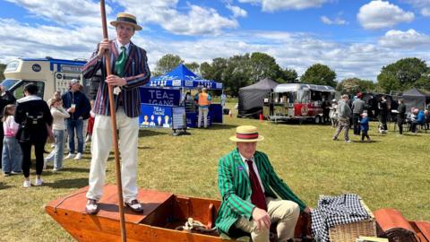 Two performers in striped blazers and straw hats pose on a small wooden boat display at an outdoor festival, with food stalls, tents and visitors spread across a grassy field behind them.