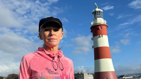 Nicky Putman is wearing a pink running jumper and a black cap on a bright sunny day in Plymouth. She is stood on grass looking straight at the camera. There is a lighthouse and the sea in the background. 