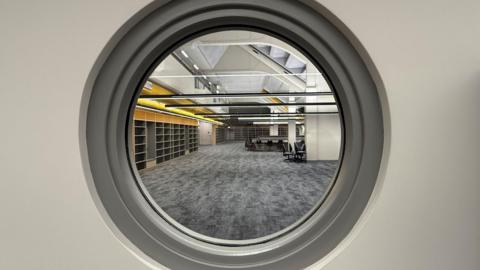 View through a grey rimmed porthole into a long library room with grey marl carpet and, to the left, shelves waiting for books to arrive. Above the shelves the wall and part of the ceiling is yellow. There are low hanging strip lights in rows and black furniture slightly too far away to see properly.