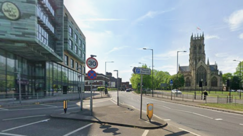 A Google Street View image of the junction of Church Way and High Fisher Gate. To the right of the picture is a large church with a St George Flag flying from the top of the tower.