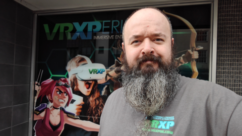 A close-up of Marc Edden, who has a long black and grey beard, smiling to camera outside his virtual experience store with a poster of cartoon characters wearing VR headsets